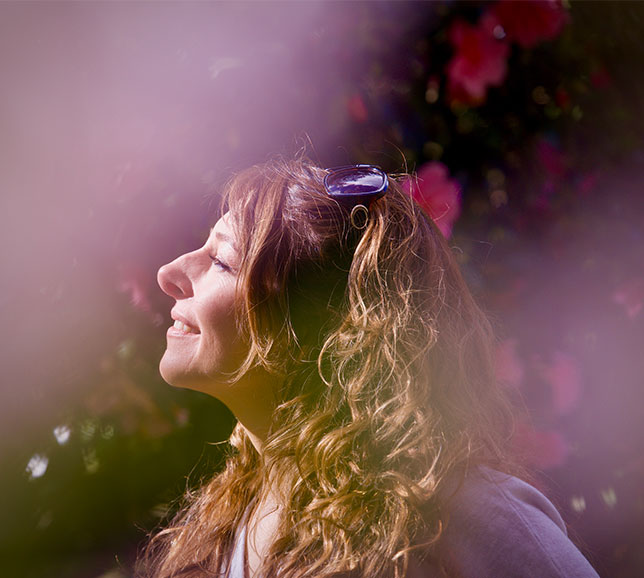 Woman with curly hair and sunglasses on head, smiling with eyes closed, surrounded by dreamy pink and purple blur, flowers in background.