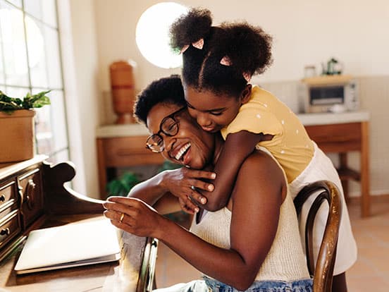 Smiling woman sitting at a desk as a young girl hugs her from behind