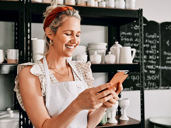 Smiling woman wearing an apron and headband checks her phone in a pottery shop, shelves of white ceramics behind her.