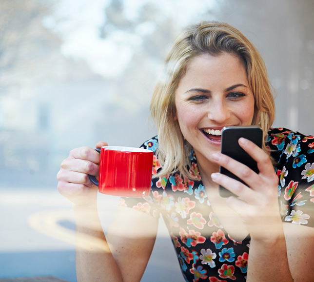 Woman drinking out of a red mug and smiling at her smart phone