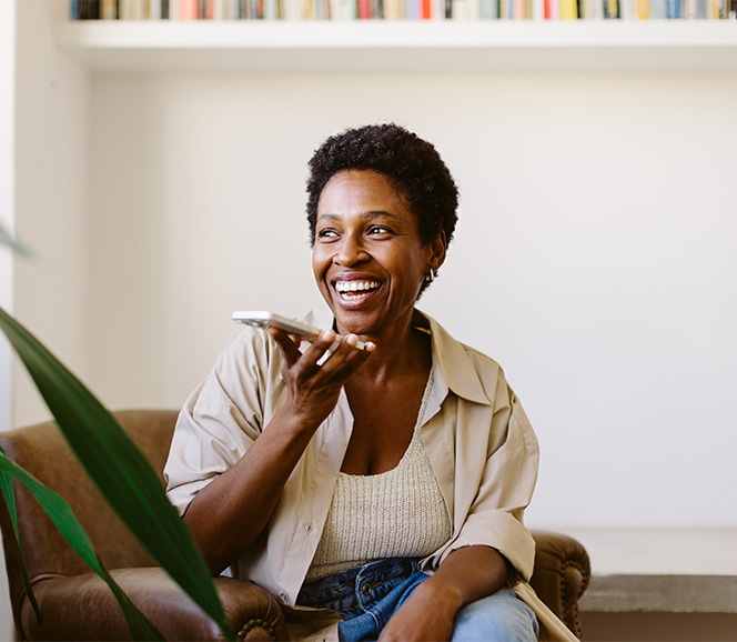 Smiling woman sitting in an armchair, speaking into a smartphone. Background features a white wall with a shelf of books.