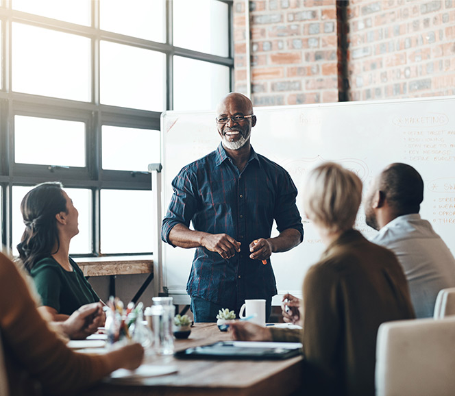 Man leading a meeting, standing by a whiteboard, engaging with four colleagues