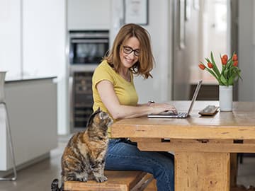 Woman in yellow shirt using a laptop at a wooden table, while a cat sits nearby. A vase of tulips is in the background.