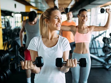 People exercising with dumbbells in a gym, smiling and engaging in a group workout session in bright, modern surroundings.