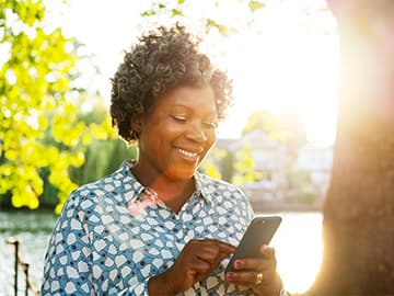 Woman standing by a sunlit lake, smiling and using a smartphone, wearing a patterned blouse. Bright greenery in the background.