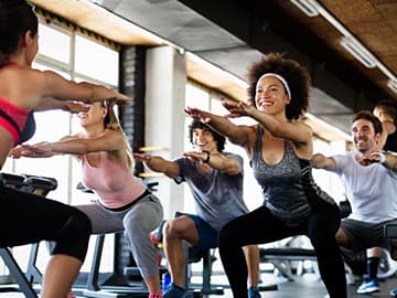 People in workout attire doing squats in a gym class, smiling and focused, with exercise equipment in the background.