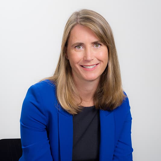 A woman with long light brown hair wearing a blue blazer and black top smiles at the camera against a plain white background.