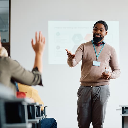 Smiling presenter reaches toward a raised attendee's hand while giving a presentation in front of a projected slide.