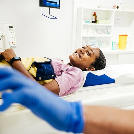 Patient smiling while lying on a medical imaging machine, wearing a protective apron. A healthcare professional's gloved hand is in the foreground.