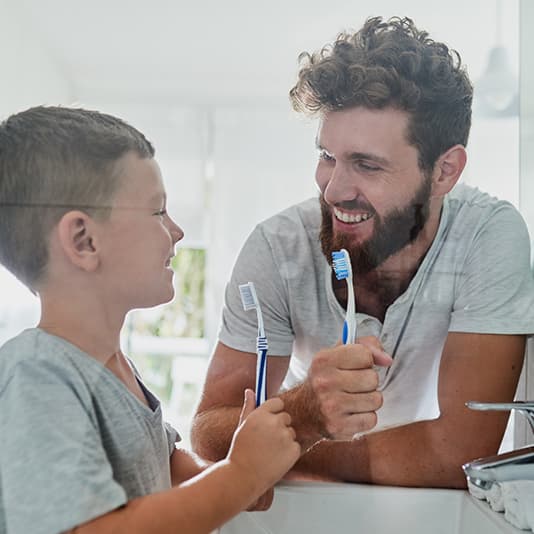 A man and a boy smiling at each other in a bathroom, holding toothbrushes, ready to brush their teeth together.