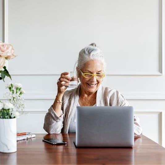 Smiling woman with yellow glasses holds a drink, working on a laptop at a wooden table with flowers and a smartphone.