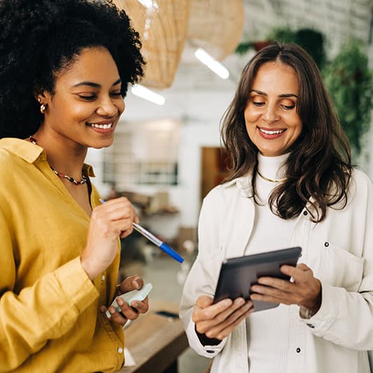 Two women smiling and discussing something on a tablet in a bright, modern workspace. One holds a pen and notes, the other holds the tablet.