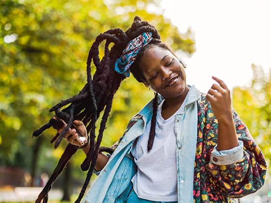 Person joyfully dancing outdoors, wearing a colourful jacket and headscarf, with long dreadlocks and a phone in hand, surrounded by greenery.