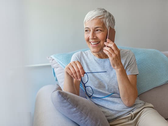Smiling woman with short gray hair sitting on a sofa, talking on her phone, and holding reading glasses