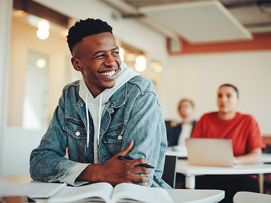 Smiling student in a denim jacket at a desk with open book and pen, classmates and laptops blurred behind.