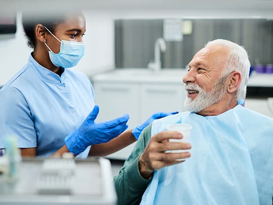 Dental hygienist with mask and gloves talks to smiling older man holding a cup in a dental office.