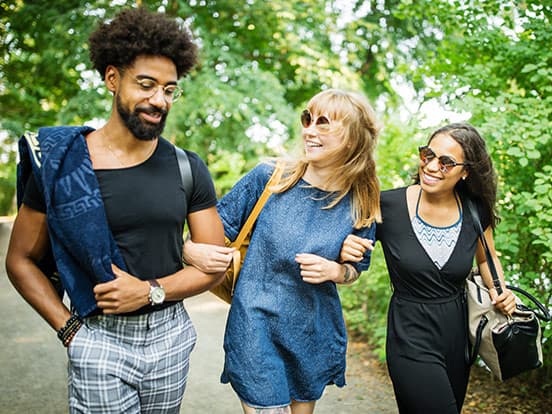 Three friends walking arm-in-arm on a tree-lined path, smiling and enjoying a sunny day, wearing casual summer outfits and sunglasses.