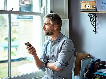 Person with a beard holding a phone, looking out the window. They're sitting in a cozy, well-lit room with shelves and office supplies.