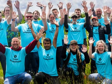A group of smiling people in blue shirts with "Team Simplyhealth" text, raising hands outdoors on a sunny day.