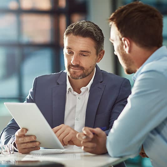 Two men in a discussion at a bright office, one holding a tablet. Both wear suits, focusing on the screen.