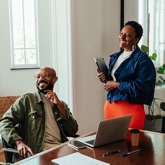 Two colleagues smiling in a bright office. One sits at a desk with a laptop, while the other stands holding a tablet.
