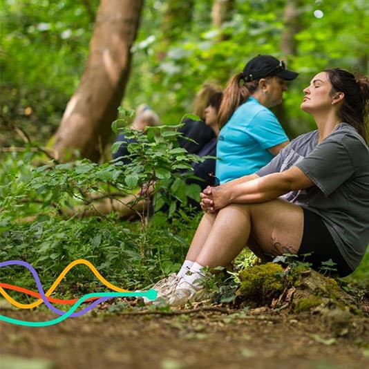 A woman sits peacefully in a lush forest, eyes closed, surrounded by others in casual attire, enjoying the natural setting.