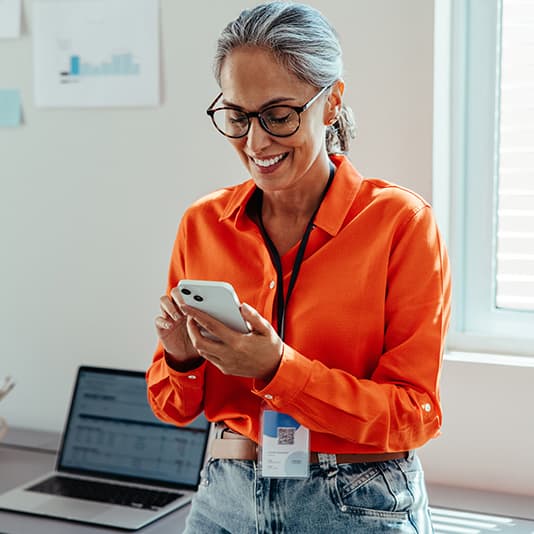 A woman in an orange shirt and glasses smiles while looking at her phone, standing in an office with a laptop and documents in the background.