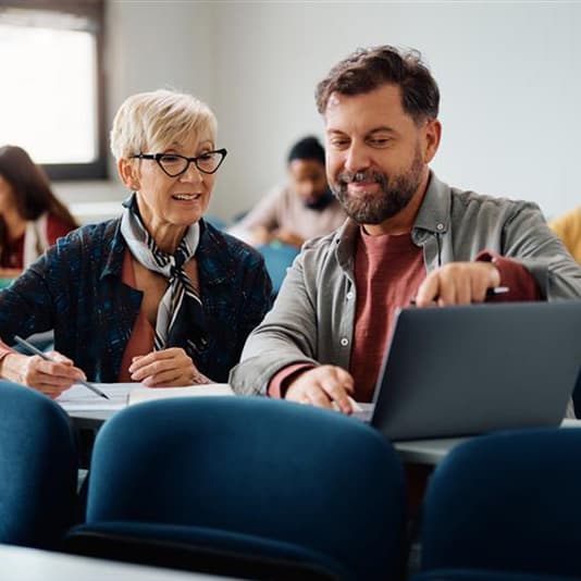 Smiling older woman and bearded man working together on a laptop and notes in a classroom.