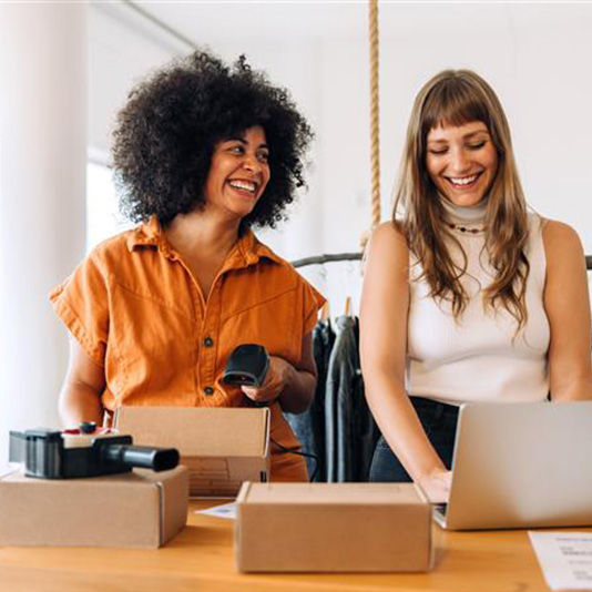 Two smiling coworkers packing shipping boxes at a table—one holding a barcode scanner, the other typing on a laptop.
