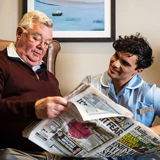 An elderly man in a chair reads a newspaper with a smiling caregiver in a light blue uniform beside him. A painting hangs on the wall behind them.