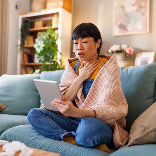 A woman sitting on a soaf holding a tablet, wrapped in a blanket, touching her throat with a concerned expression.