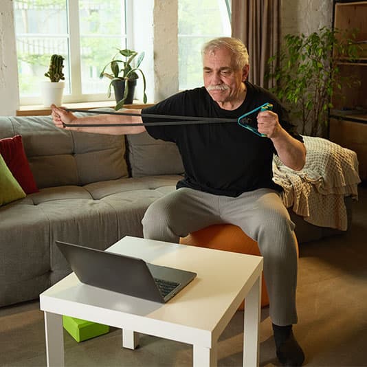 A man sits on an exercise ball, stretching a resistance band while looking at a laptop in a cozy living room with plants and a couch.