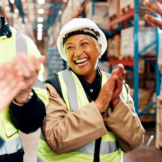 A woman in a hard hat and high-visibility vest claps and smiles in a warehouse, surrounded by colleagues.