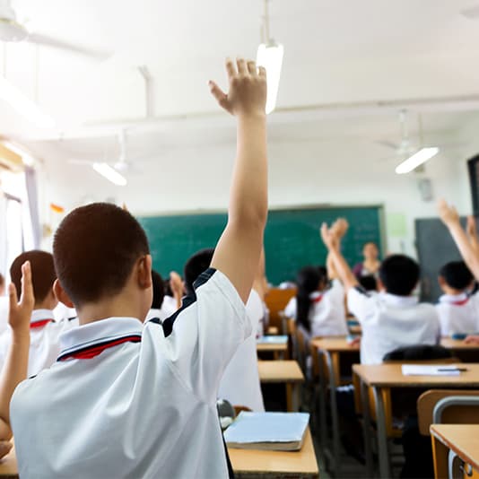 Students in a classroom with hands raised, wearing white uniforms, facing a teacher at the front near a blackboard.