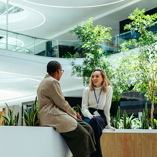 Two people sitting and talking in a modern, bright indoor space with plants and stylish architecture in the background.