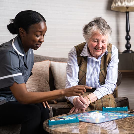 Caregiver and elderly woman playing Scrabble at a table. They are smiling and enjoying the activity together in a cozy room.
