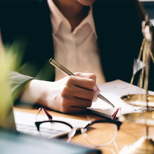 A person in formal attire writing with a pen at a desk, near glasses, a laptop, and a balanced scale.