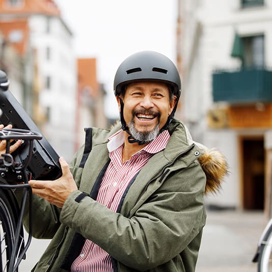 Smiling man wearing a helmet and green jacket, holding a bike component in a city street setting.
