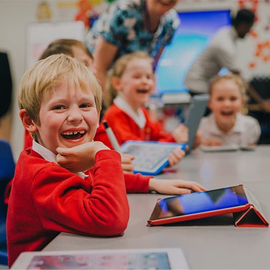 Children in a classroom, smiling and using tablets at their desks. A teacher is in the background.