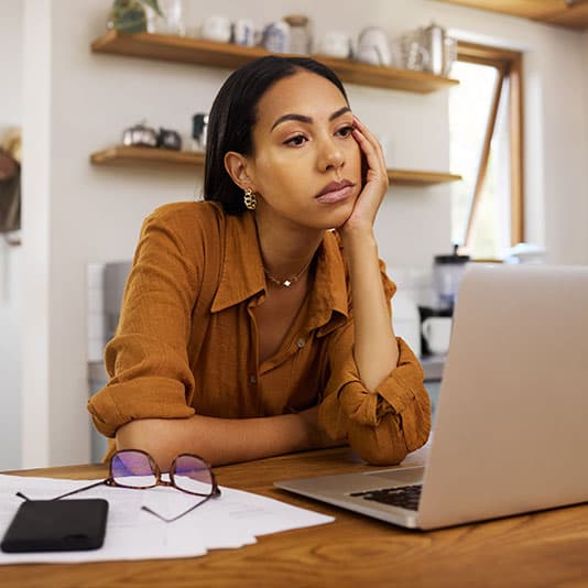 A woman in a brown shirt looks thoughtful while sitting at a desk with a laptop, phone, papers, and glasses in a cozy, well-lit kitchen.