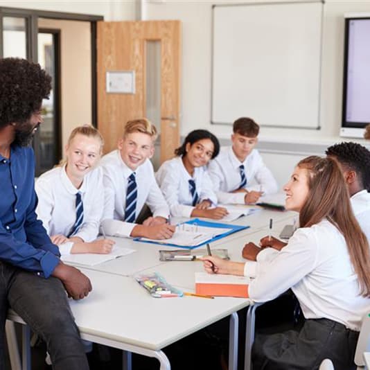 Teacher and students in school uniforms sit around a table, engaged in discussion, with papers and pens in a classroom setting.