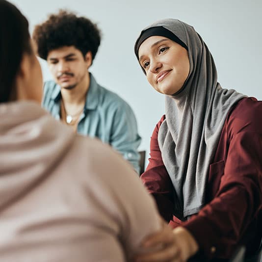 Three people are engaged in a conversation. A woman in a hijab is smiling and listening attentively to another person in the foreground.