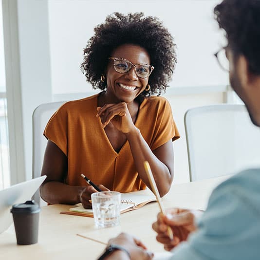 A person in glasses and an orange blouse is smiling, seated at a table with a notebook, pen, and glass of water, engaging in a discussion.