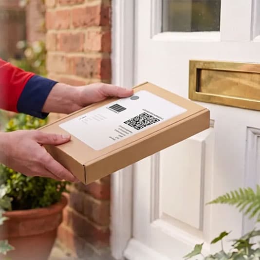 A person in a red and blue uniform delivers a flat cardboard package to a white door with a gold mail slot.