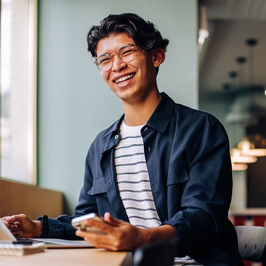 Smiling young person with glasses holding a smartphone, sitting at a table in a well-lit room with large windows.