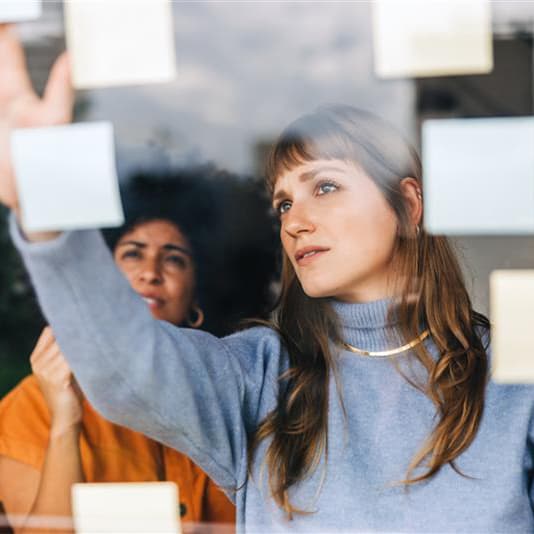 Two women are seen through a glass window, arranging colorful sticky notes. The woman in front wears a blue sweater and reaches towards the notes.