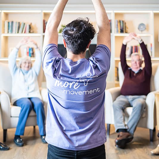 Instructor leading a seated exercise class with seniors in a cozy room, featuring a wall of bookshelves in the background.