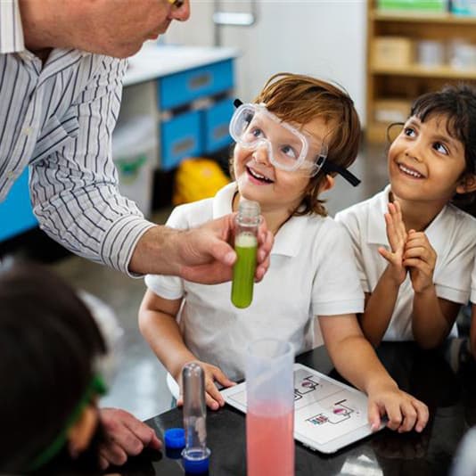 Teacher shows a test tube with green liquid to smiling children (one wearing safety goggles) during a classroom science experiment.