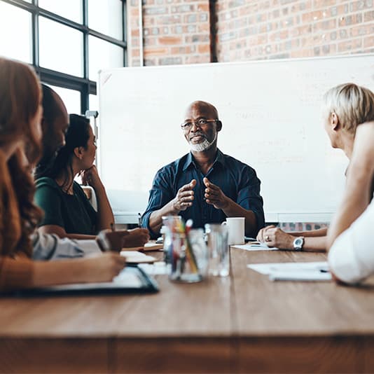A person leads a discussion at a table with five others, in front of a whiteboard. They appear engaged in a collaborative meeting.