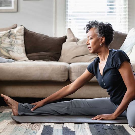 Woman stretching on a yoga mat in a living room, touching her foot with a calm expression. Couch and decorative pillows in the background.